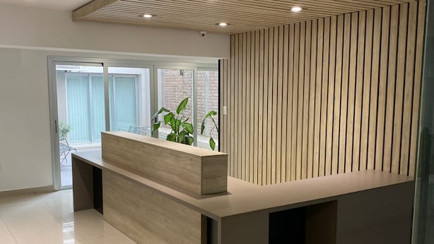 A reception desk with a modern design, a large window with a view of a brick building, and a potted plant on the desk. - HOMEFULL  APARTAMENTOS (Salta)