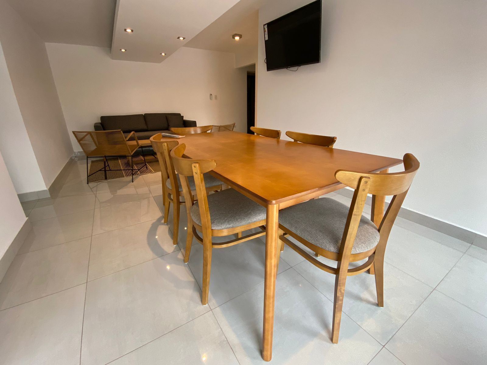 Un comedor moderno con una mesa y sillas de madera, un televisor de pantalla plana en la pared y un sofá al fondo.