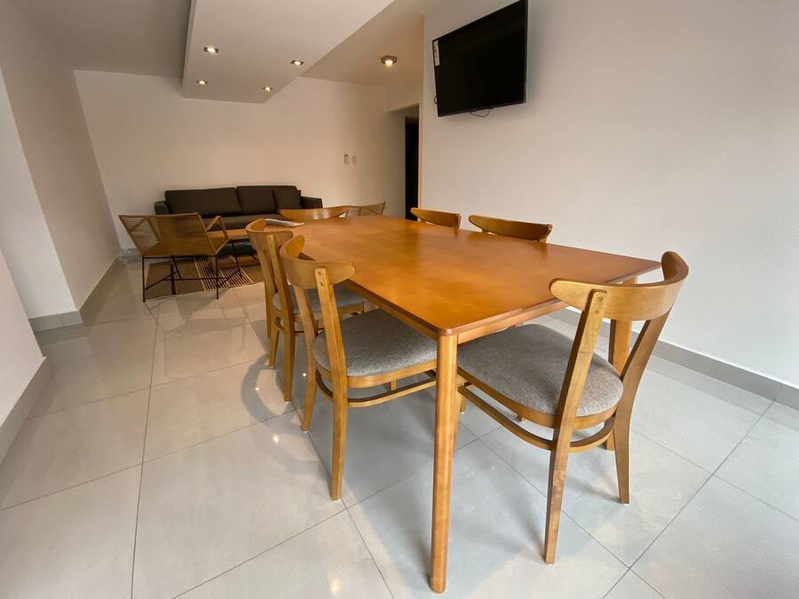 A modern dining area with a wooden table and chairs, a flat-screen TV mounted on the wall, and a sofa in the background.