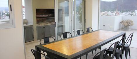 A dining area with a long wooden table and black chairs, a sliding glass door leading to a kitchen, and a view of a cityscape through a large window.