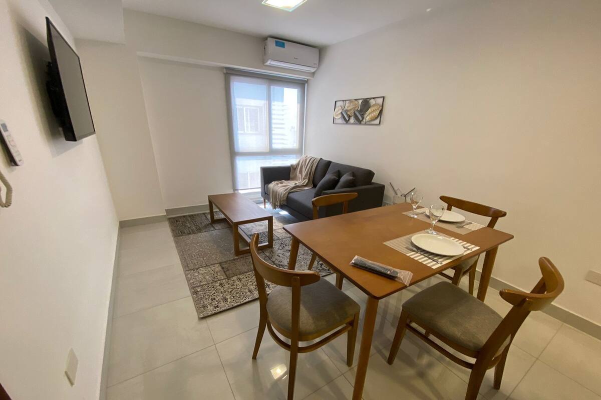 A dining area with a wooden table and four chairs, a small rug, and a sofa in the background.