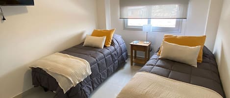 Habitación con dos camas, un televisor de pantalla plana montado en la pared, una ventana con persianas y una lámpara en el techo.