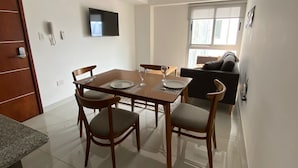 A dining area with a table set for two, two chairs, a flat-screen TV mounted on the wall, and a window providing natural light. - HOMEFULL  APARTAMENTOS (Salta)