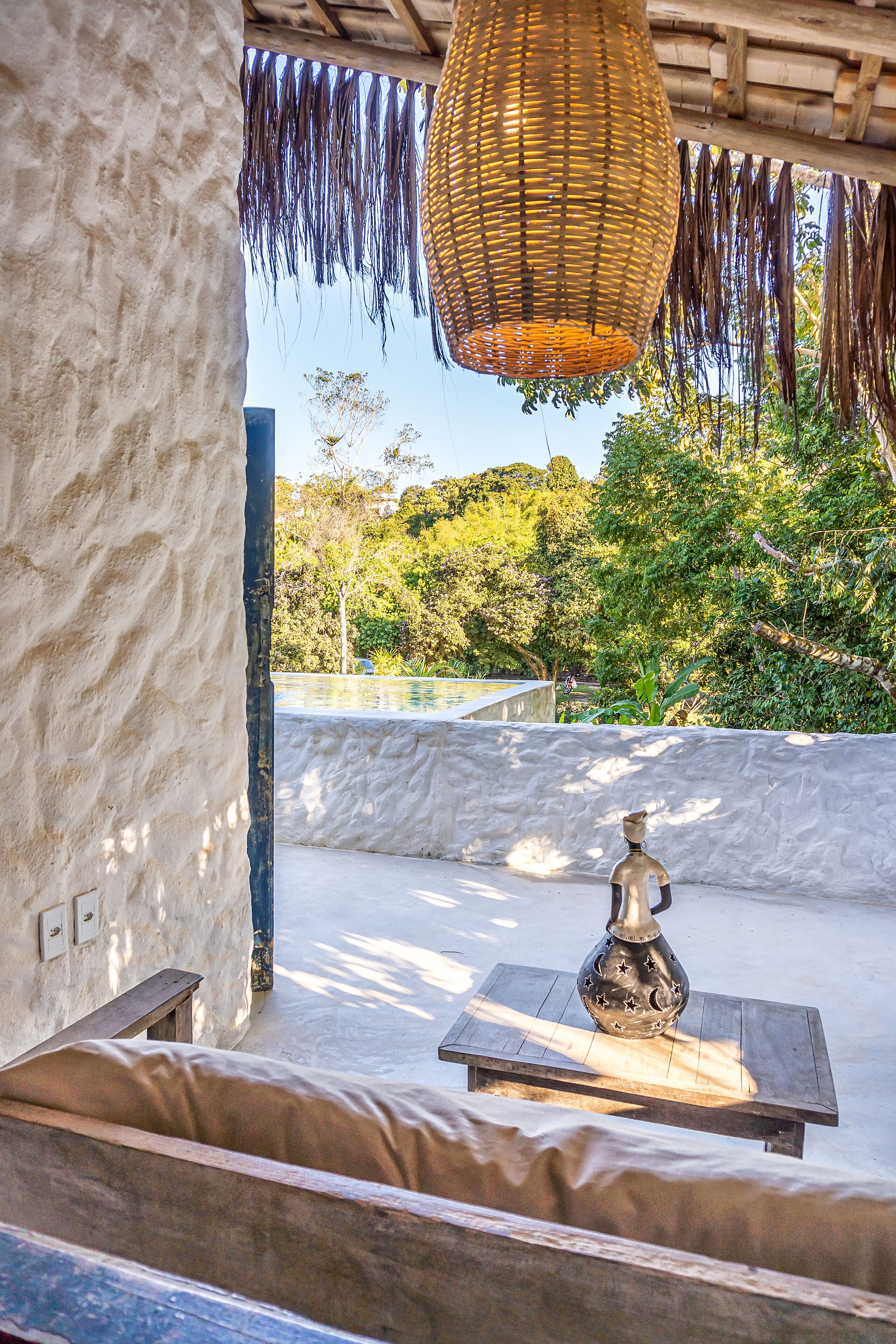A room with a stone wall, a woven hanging light fixture, a wooden table with a decorative vase, and a view of a pool and greenery outside.