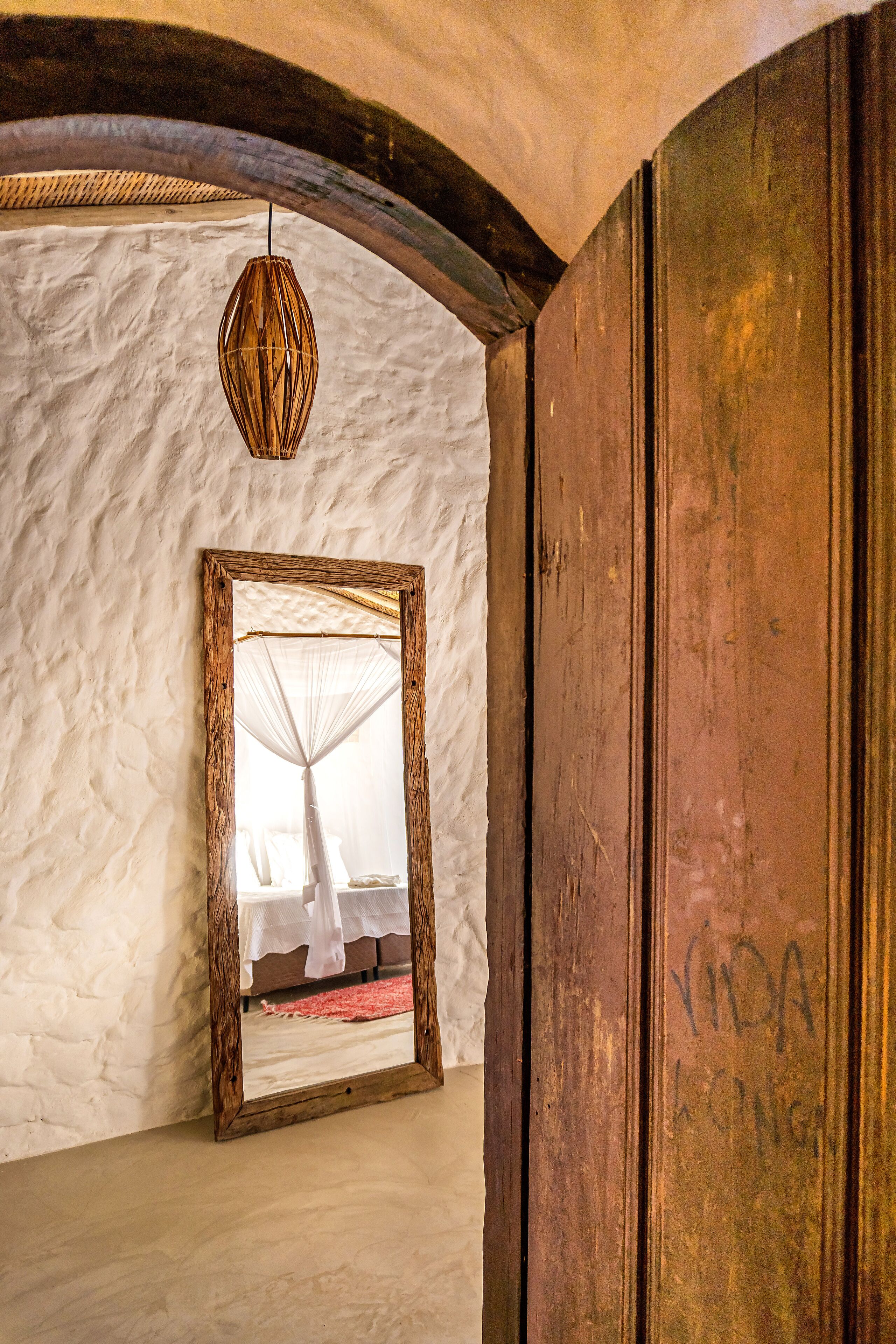 Ein Zimmer mit einer Wand mit Strukturmuster, einem spiegelnden, holzrahmenartigen Spiegel, der ein Bett mit weißem Baldachin zeigt, und einer hängenden Leuchte.