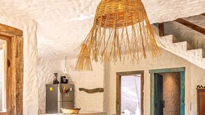 Un interno rustico con soffitto in pietra, un lampadario intrecciato, un angolo cottura con frigorifero e una zona giorno accogliente con una panca e sedie.