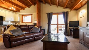 A living room with a leather sofa, a coffee table, wooden beams on the ceiling, a kitchen in the background, and a TV on a stand. - Avocet Lodge (Lincoln)