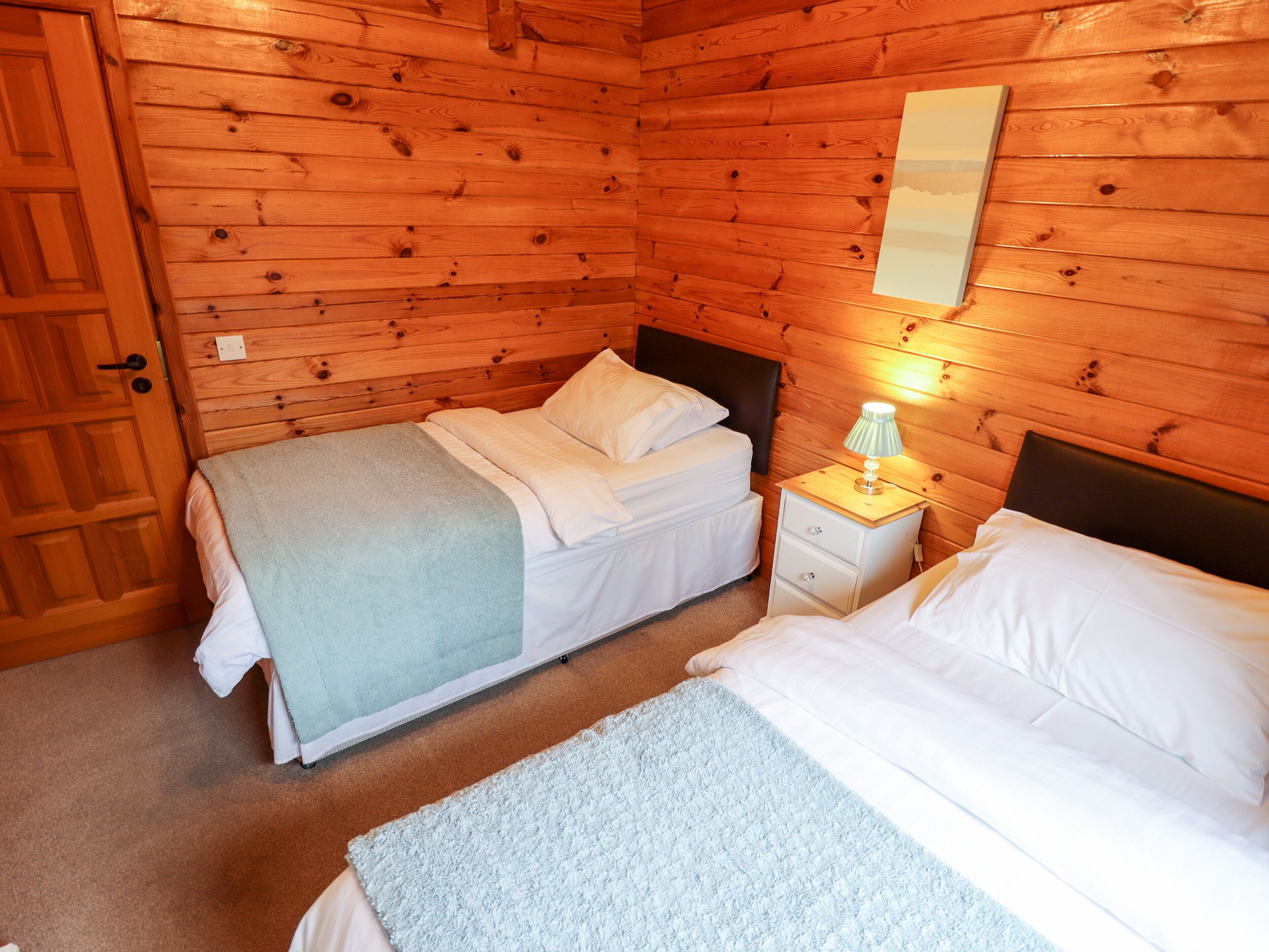 Un dormitorio de cabaña de madera con dos camas, una mesita de noche con lámpara y una puerta de madera.