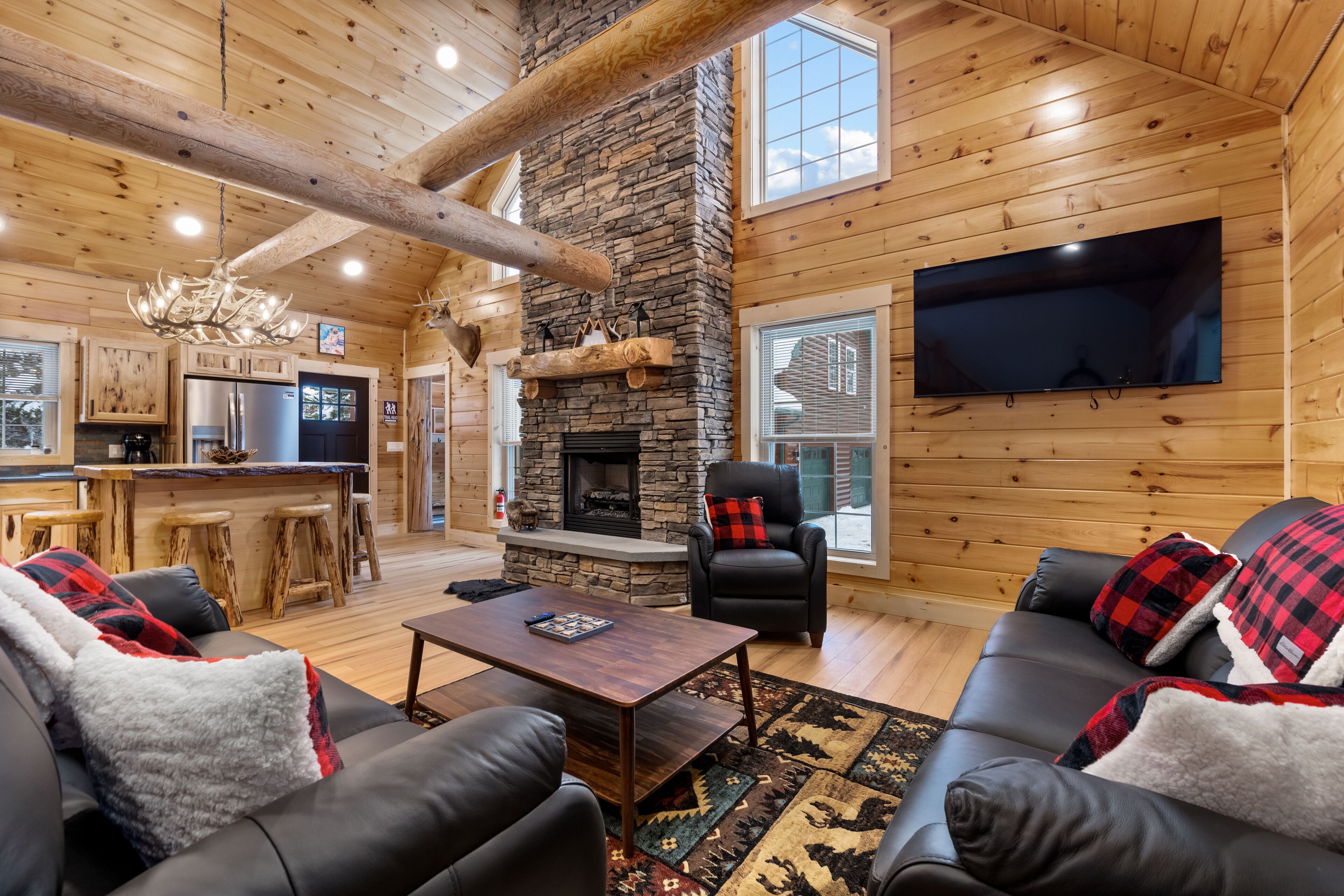 A log cabin interior with a stone fireplace, wooden walls, a flat-screen TV, leather sofas, and a bar area.