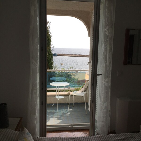 Habitación con puerta de vidrio corredera que da a un espacio exterior con mesa y sillas, vista al mar y un edificio al fondo.