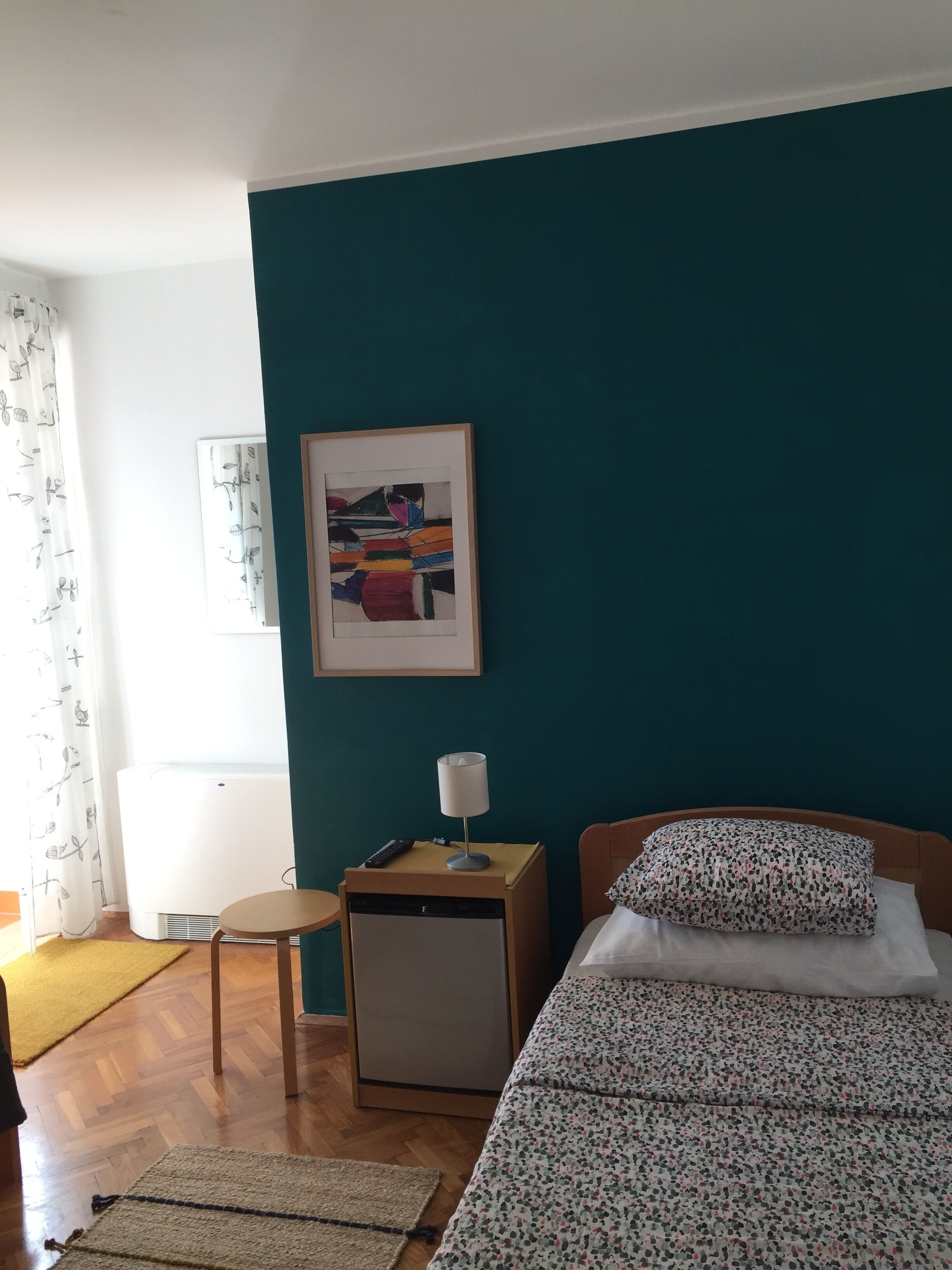 A bedroom with teal walls, a bed with a floral duvet, a bedside table with a lamp, a small cabinet, and a framed abstract painting.