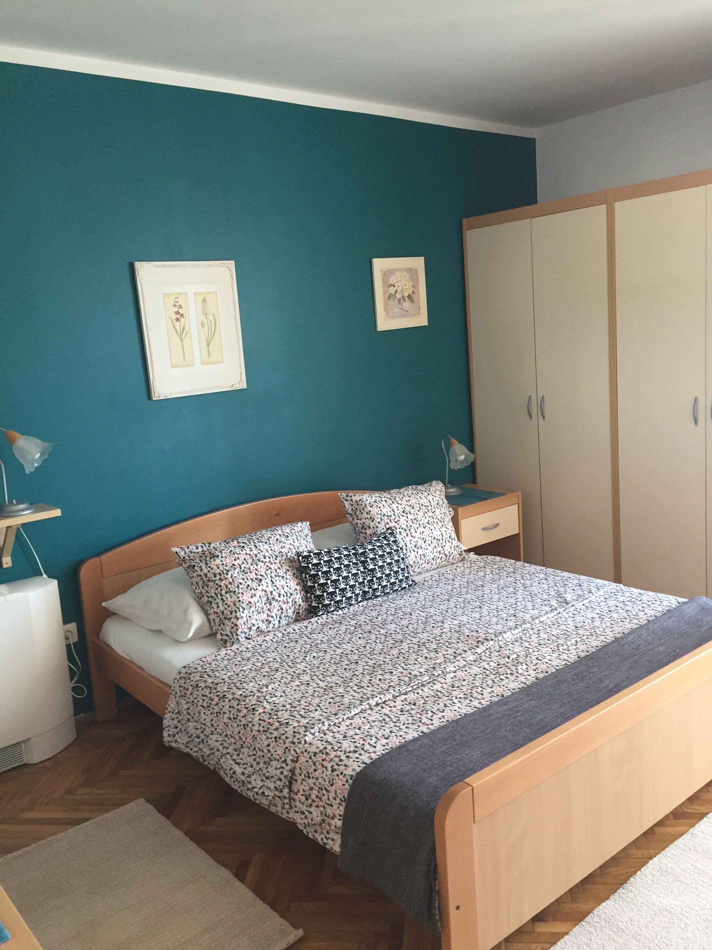 A bedroom with a bed, wooden headboard, two bedside tables with lamps, a wardrobe, and a teal wall with framed botanical prints.