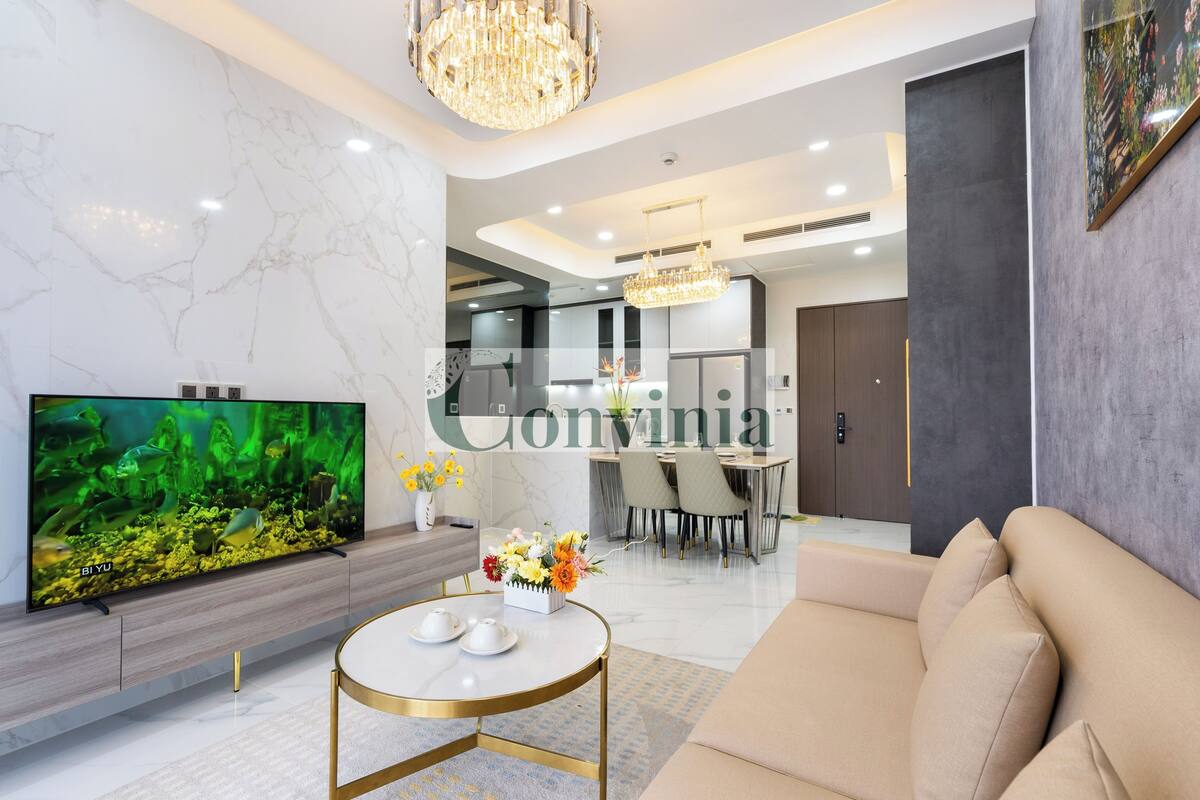 A modern living room with a large flat-screen TV, a round coffee table, a beige sofa, and a chandelier.