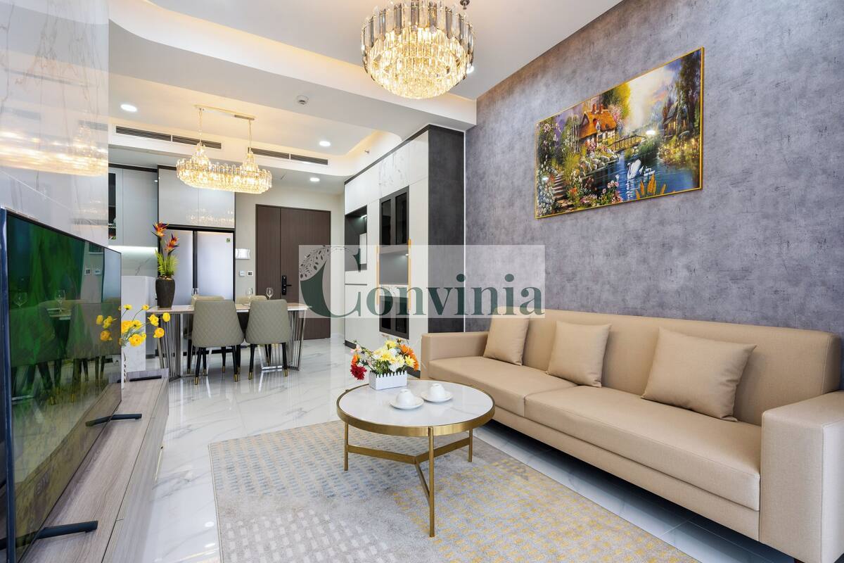 A modern living room with a beige sofa, a round coffee table, and a chandelier. There is a painting on the wall and a dining area in the background.