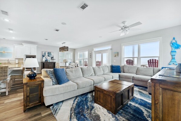 A spacious living room with a large sectional sofa, a wooden coffee table, and a blue and white rug. There is a kitchen area with white cabinets and a dining table.