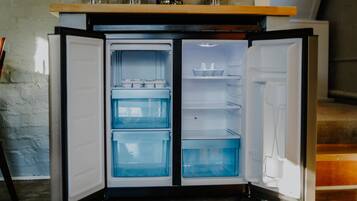 Un refrigerador compacto con varios estantes y un compartimento congelador, ubicado en una cocina con pared de ladrillo rústico y piso de madera.