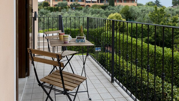A balcony with a table and chairs, a potted plant, and a view of a building and greenery. - House in the Olive Trees-For a relaxing vacation at the lake. (Lombardia)