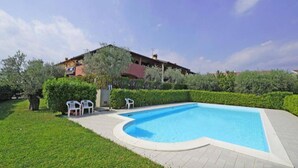 A swimming pool with a curved edge, surrounded by a paved deck. There are two white chairs near the pool's edge. - House in the Olive Trees-For a relaxing vacation at the lake. (Lombardia)