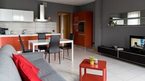 A modern living room with a grey sofa, red pillow, and a small red table with a plant. A kitchen with white cabinets and a red appliance is visible in the background. - House in the Olive Trees-For a relaxing vacation at the lake. (Lombardia)