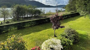 Un jardin bien entretenu, agrémenté de divers arbustes et fleurs, un mur de pierre et une vue sur un lac et des montagnes au loin.