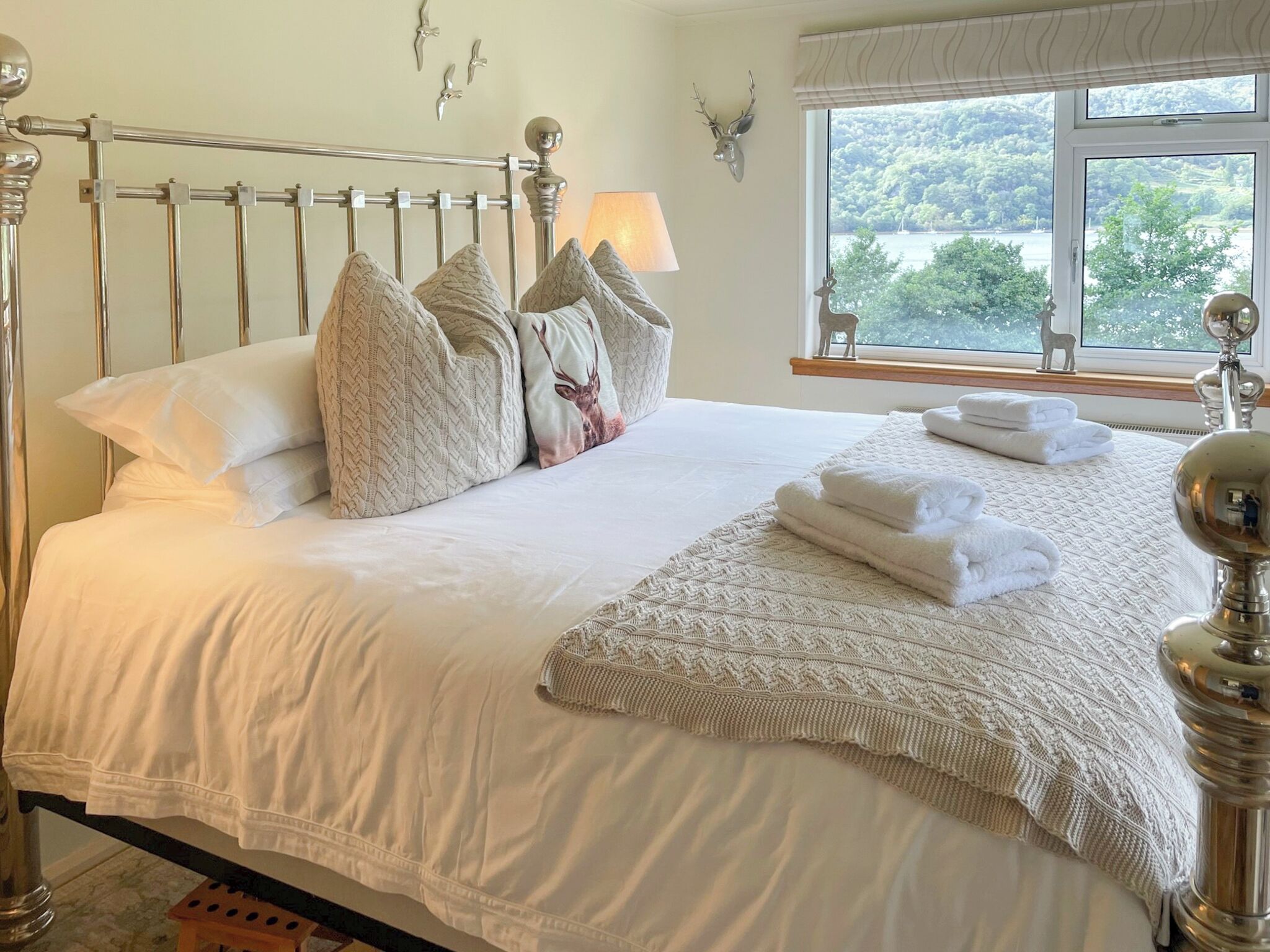Cama bem arrumada com lençóis brancos e almofadas decorativas, abajur e janela com vista para o verde.