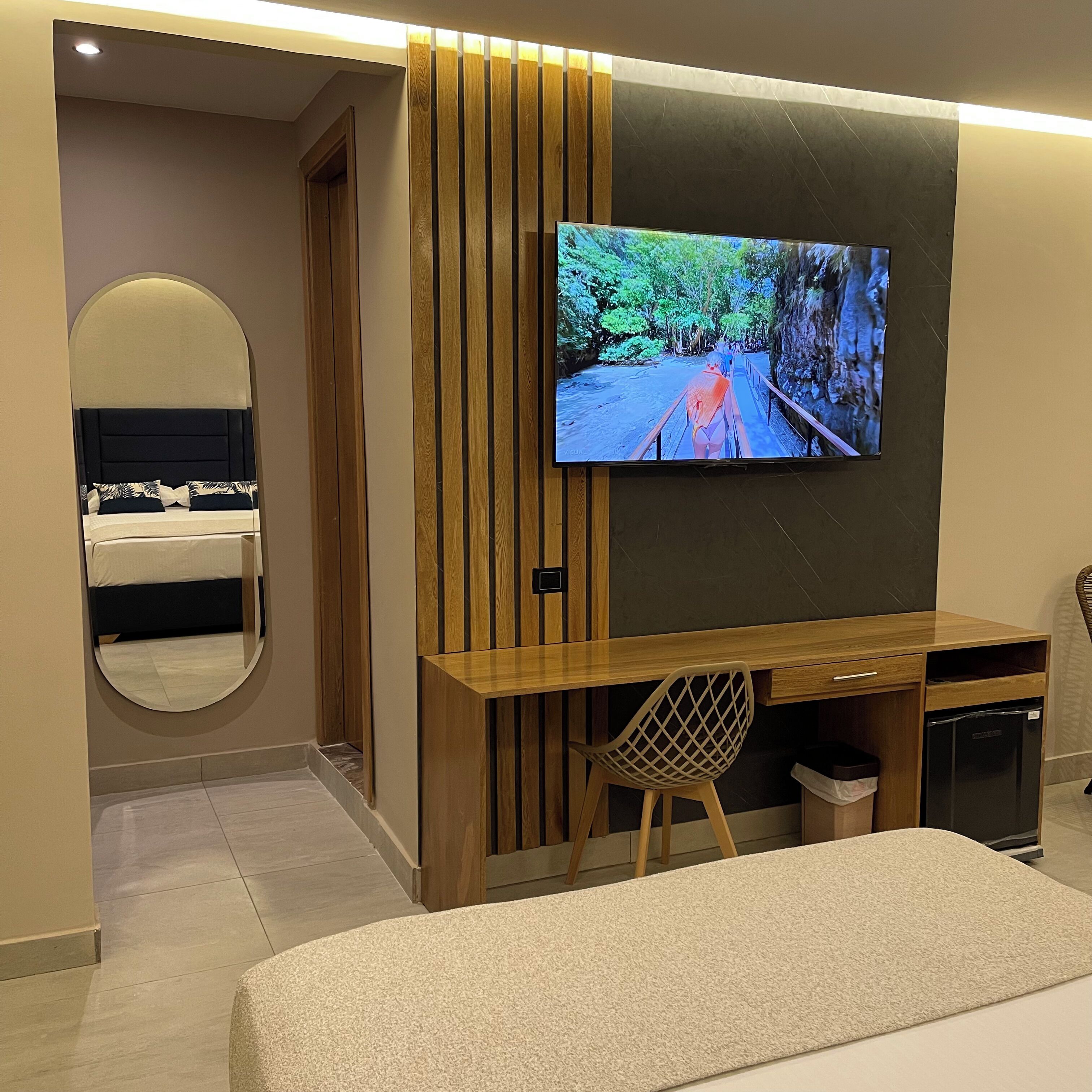 A modern hotel room with a large TV mounted on the wall, a wooden desk, a chair, a bed with a headboard, and a mirror.