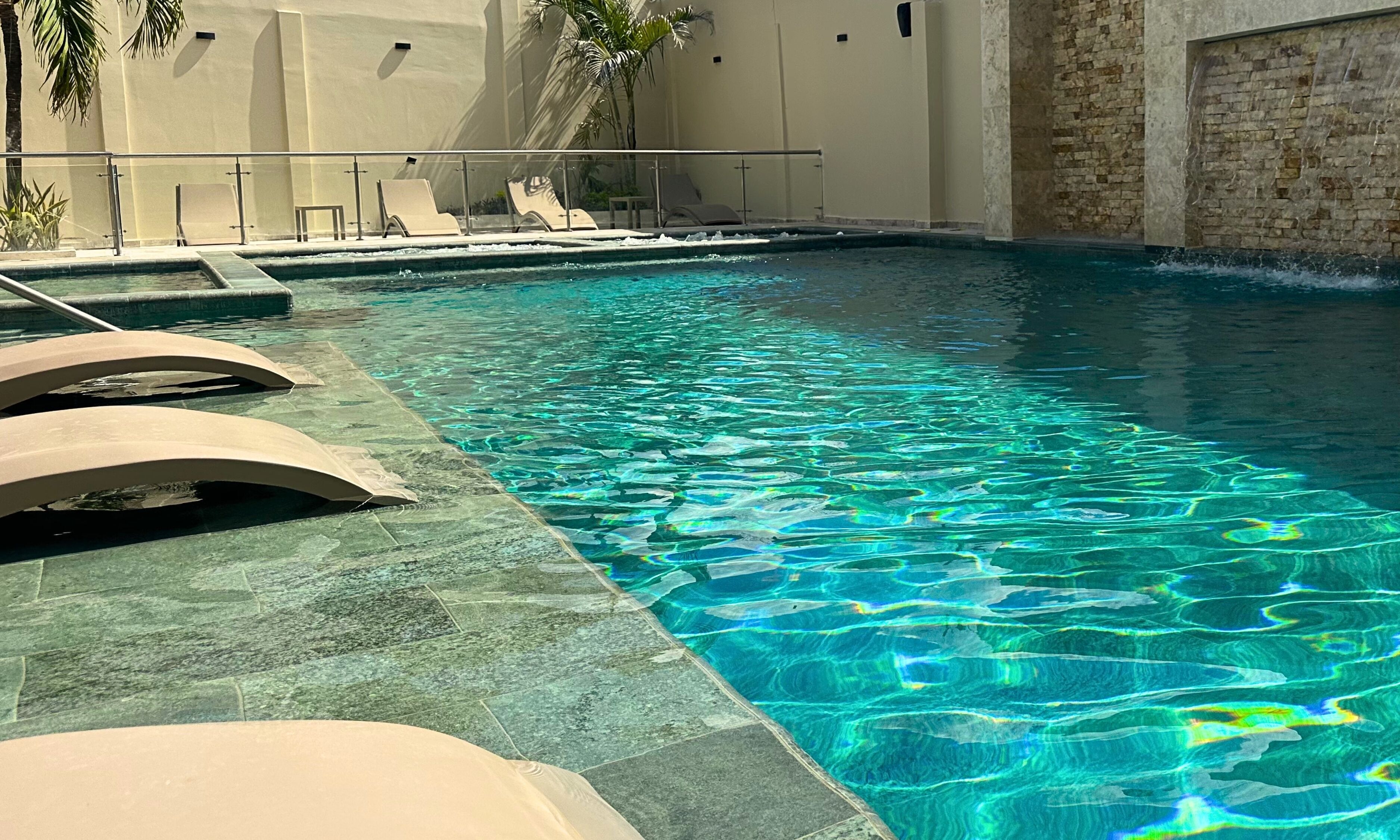 A swimming pool with clear water, lounge chairs, and a stone wall.