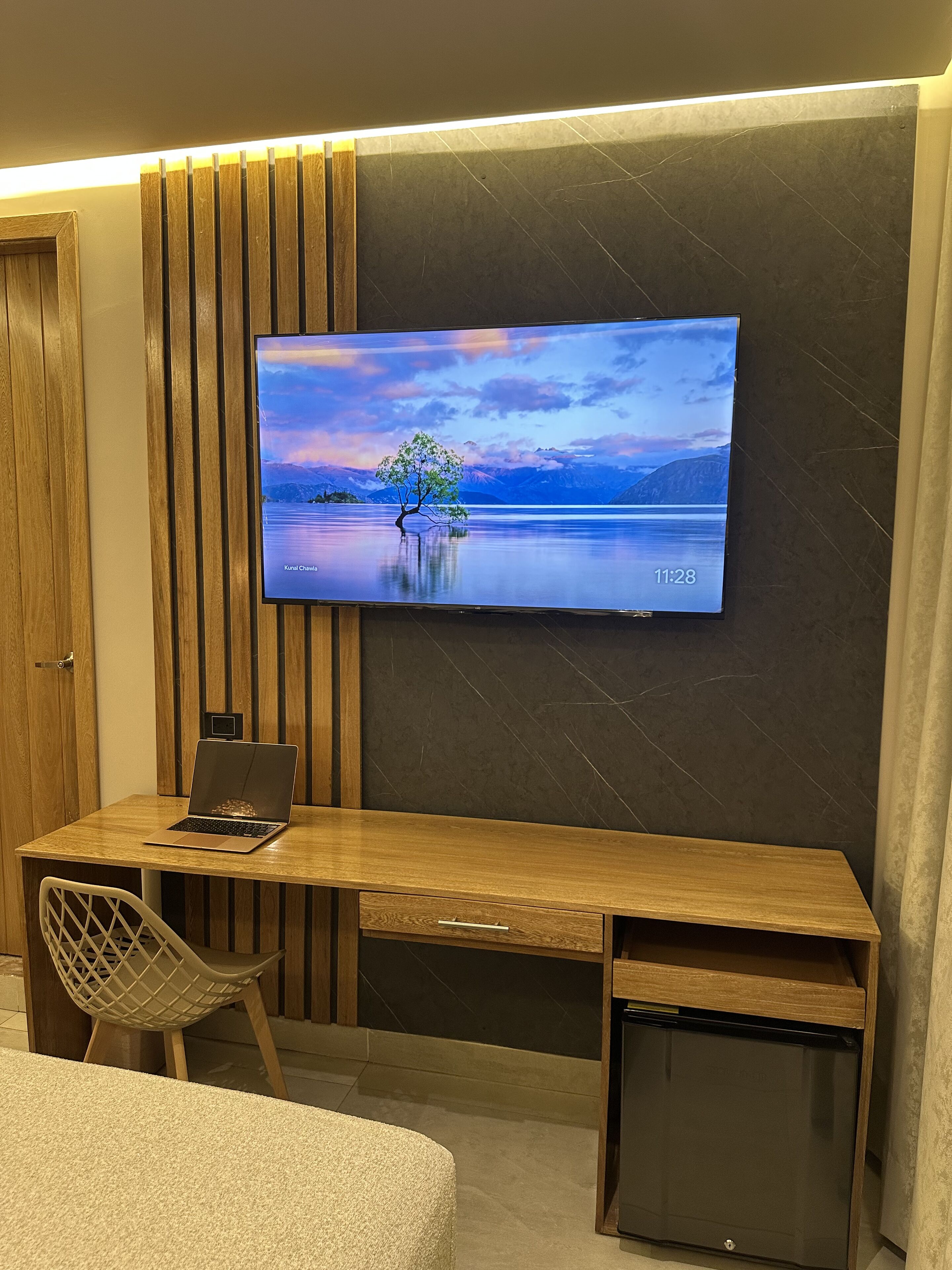 A modern workspace with a wooden desk, a laptop, and a television displaying a scenic landscape.