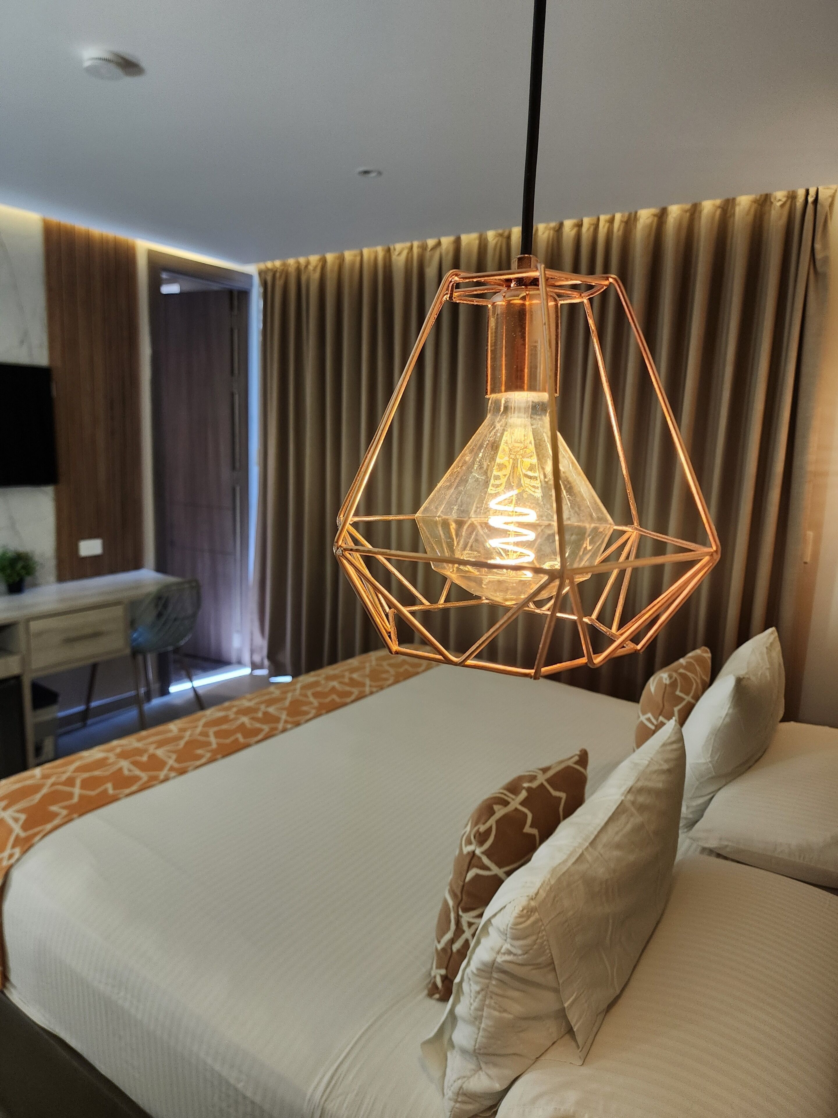 A modern hotel room with a geometric pendant light, a large bed with patterned pillows, and a view of the outside through a window with curtains.