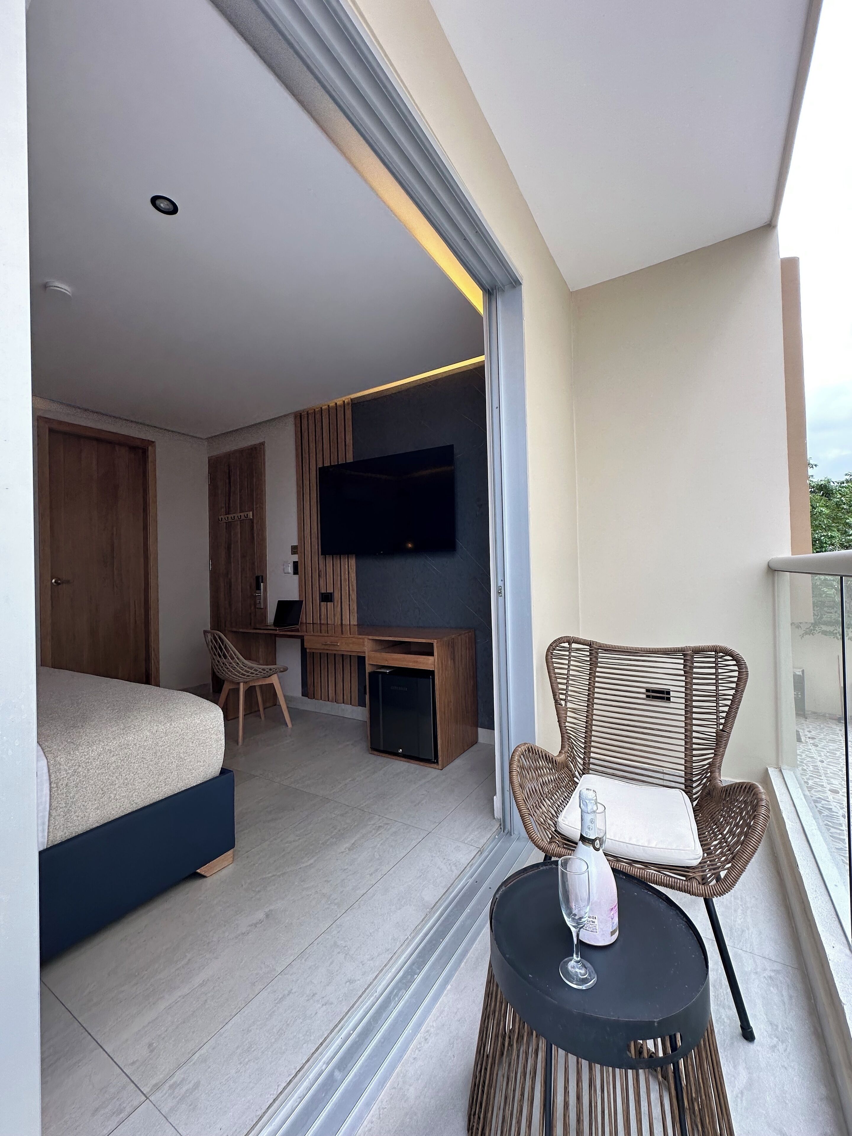 A modern hotel room with a bed, a desk, a TV, and a balcony with wicker furniture.