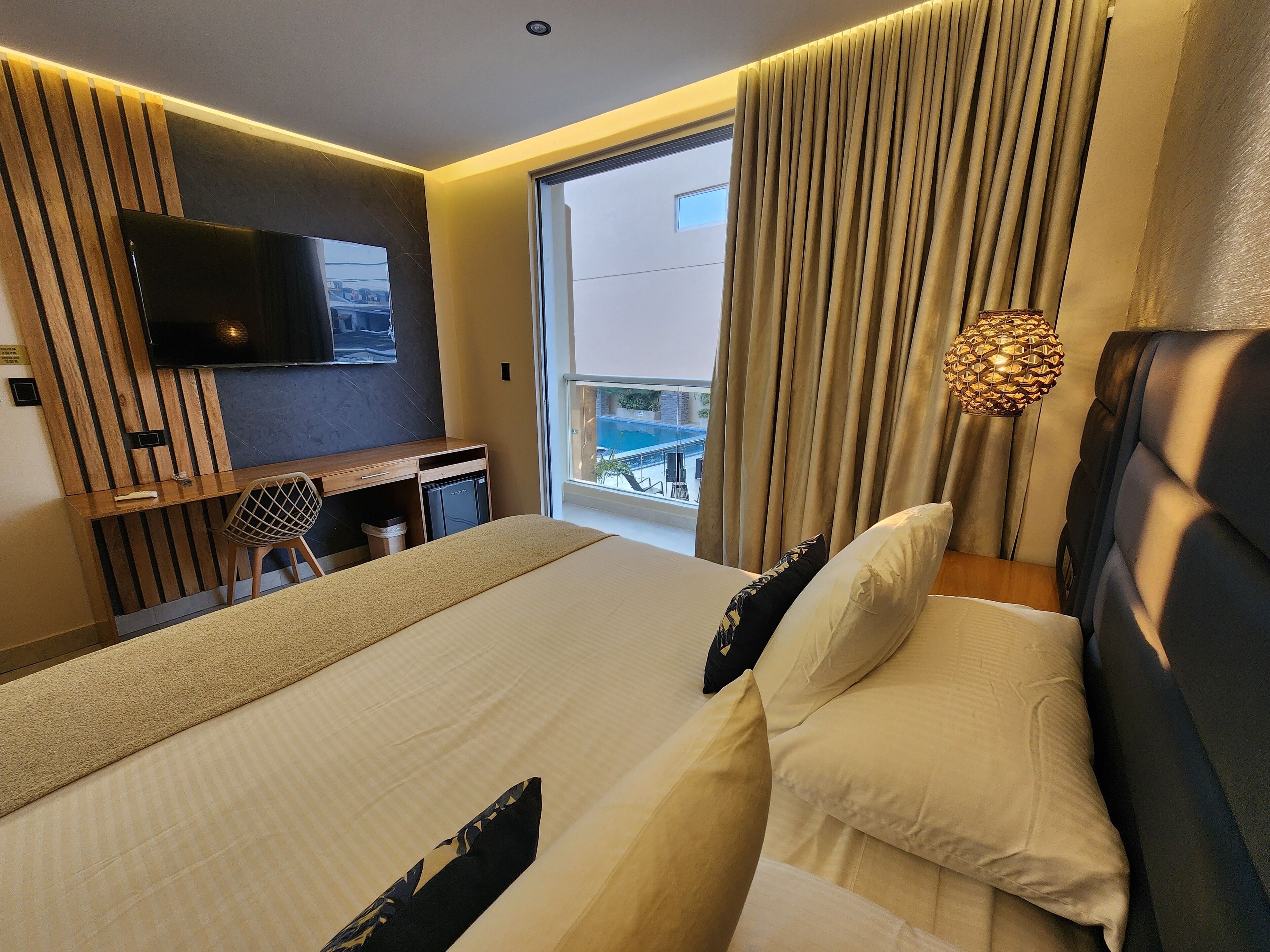 A modern hotel room with a large bed, a desk with a chair, a television, and a view of a pool.