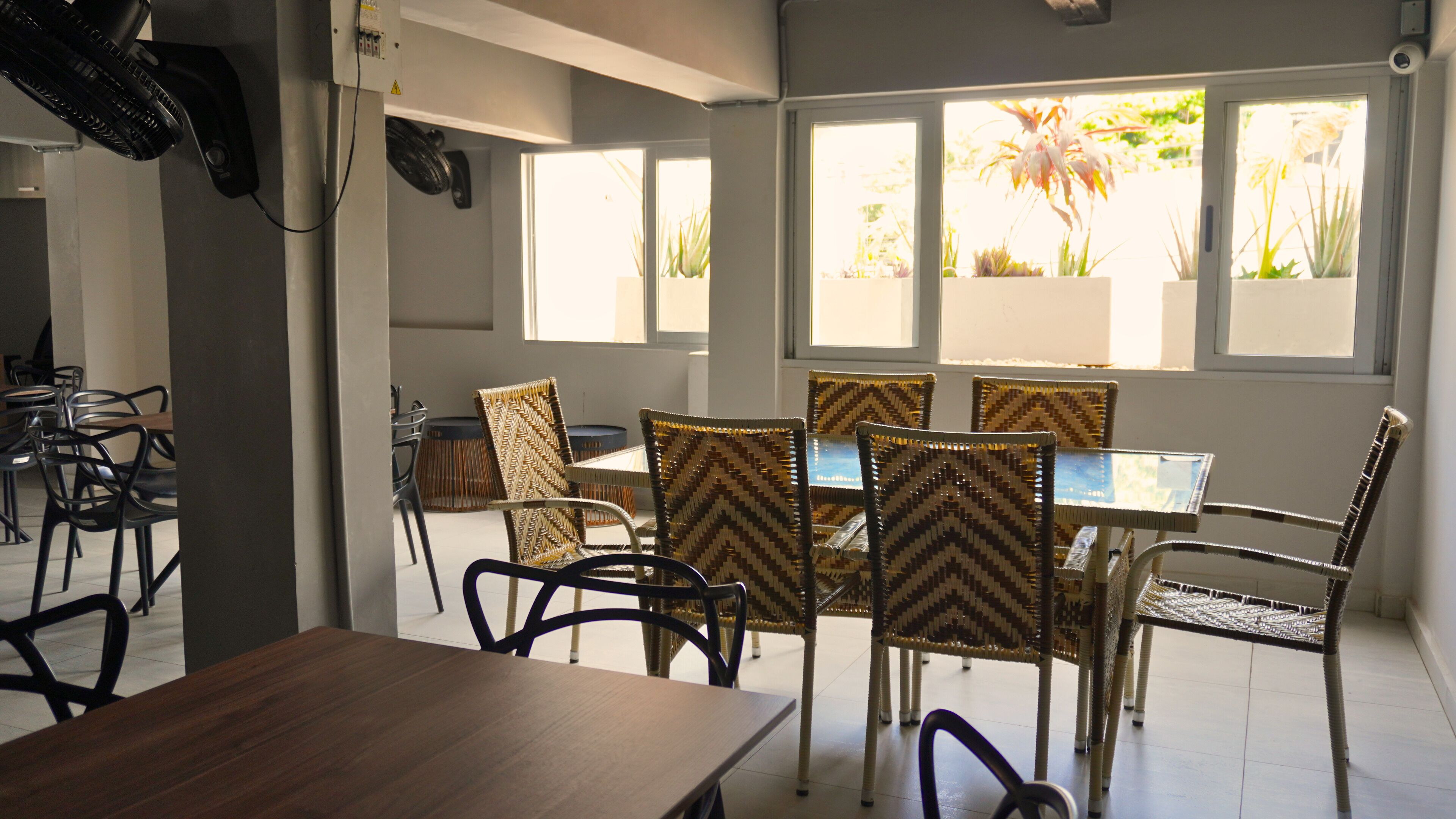 Un espace repas comprenant une table et des chaises, de grandes fenêtres donnant sur des plantes et un ventilateur de plafond.