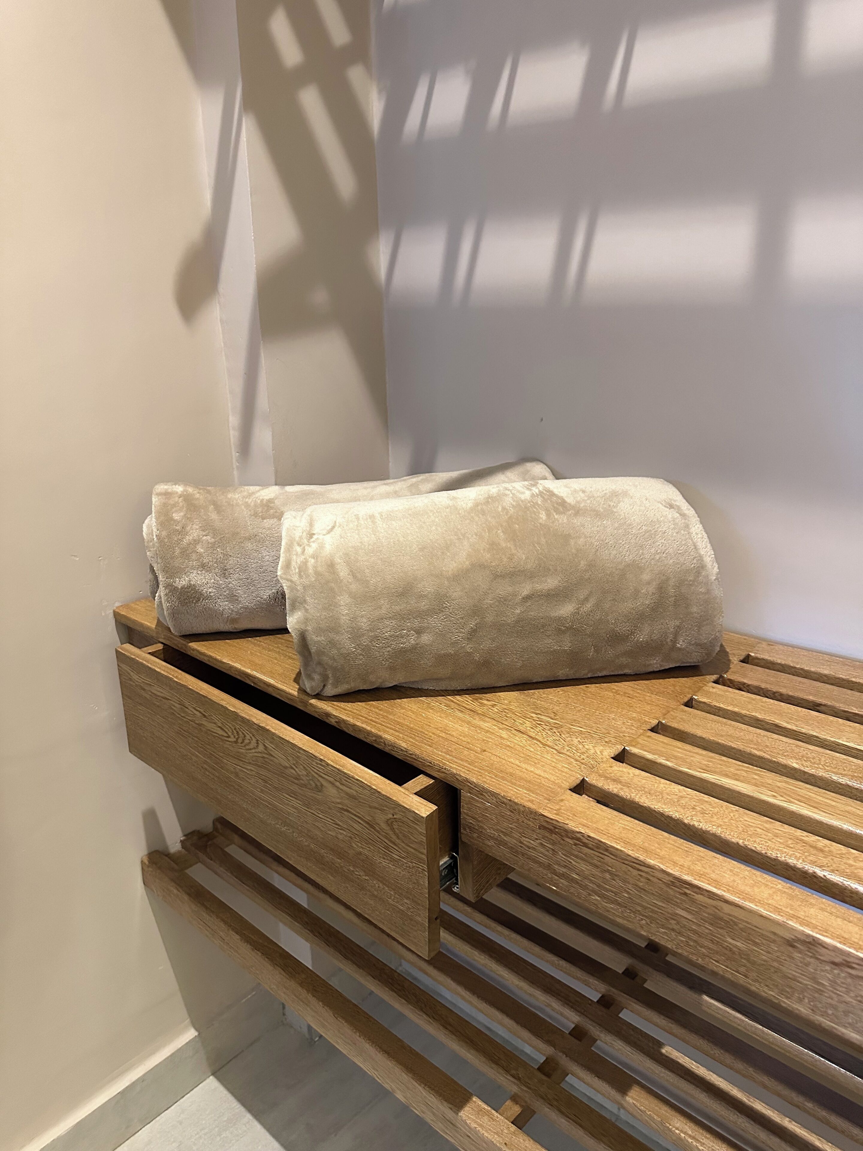A wooden bench with a built-in storage drawer, two towels, and a wall with a window casting shadows.