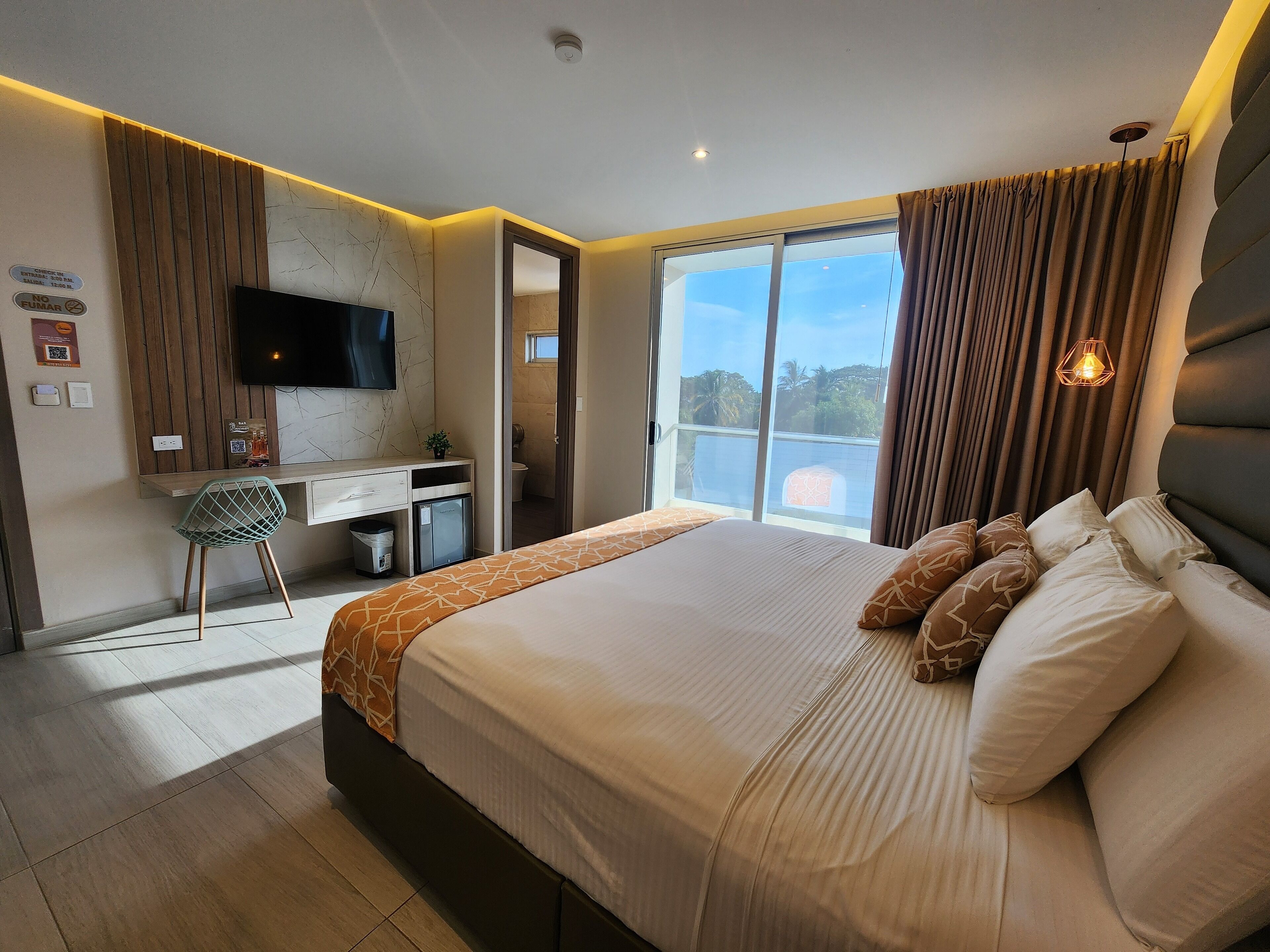 A modern hotel room with a large bed, a desk with a computer, a TV, and a balcony with a view.