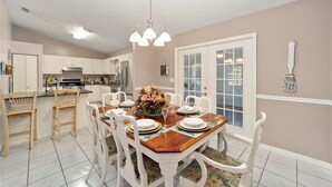 A dining room with a wooden table set for four, surrounded by white chairs, a chandelier, and a view of the kitchen. - Nellies Cottage is 4 miles from WEC-World Equestrian Center (Ocala)