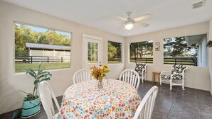 A dining area with a round table, white chairs, a ceiling fan, and large windows offering views of the outdoors. - Nellies Cottage is 4 miles from WEC-World Equestrian Center (Ocala)