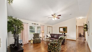 A living room with a ceiling fan, wooden flooring, floral-patterned chairs, a leather sofa, and a television on a stand. - Nellies Cottage is 4 miles from WEC-World Equestrian Center (Ocala)