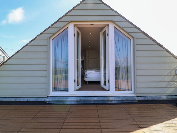 A triangular attic bedroom with a bed, white curtains, and a wooden deck.