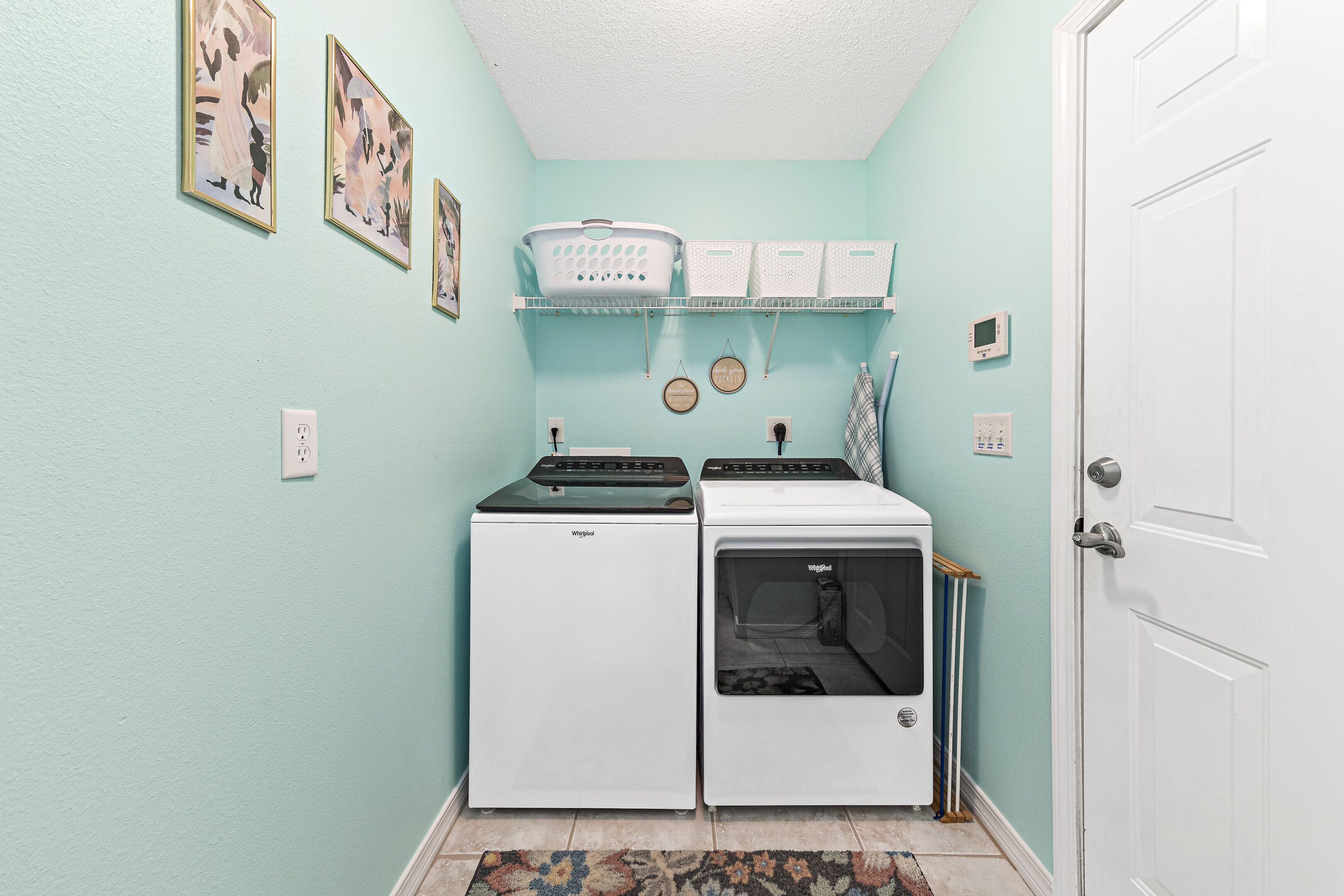 A laundry room with two front-loading washers and dryers, a shelf with laundry baskets, and a door to the right.