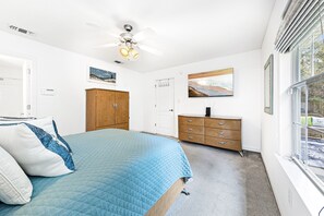 A bedroom with a bed, a ceiling fan, a wooden dresser, a door, and a window. - Lillian House (Pensacola)
