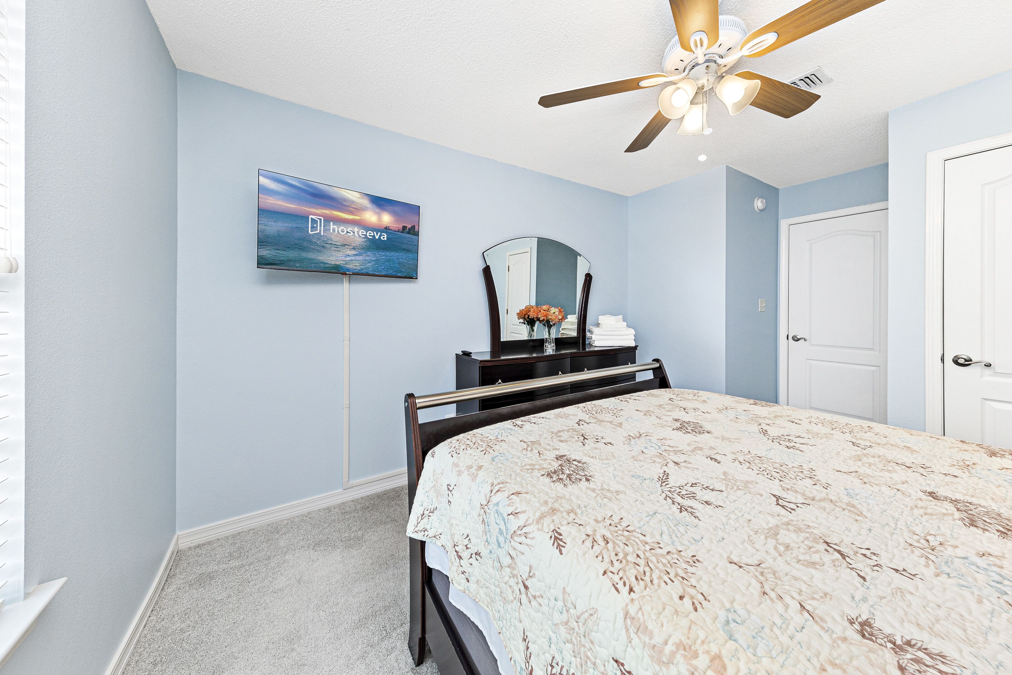 Une chambre à coucher avec un lit, un ventilateur de plafond, une télévision, une commode et une porte.