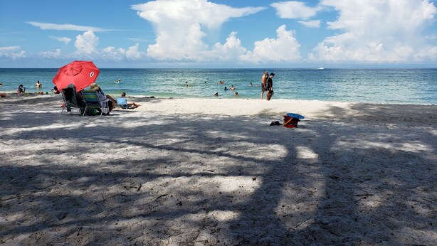A beach with people swimming and sunbathing, a red umbrella, and a boat in the distance. - Modern Condo with easy beach access in the heart of Naples Park (Naples)