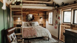 Un dormitorio acogedor con literas, techo de madera, espejo redondo, ventana con estampado floral y un escritorio pequeño.