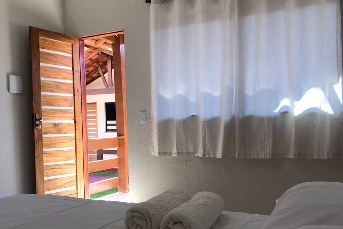 A hotel room with a bed, two rolled towels, a wooden door, and a window with curtains.