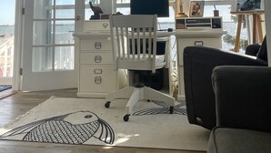 Ett rymligt rum med ett skrivbord, en stol och en dator, stora fönster med utsikt över palmer och en takfläkt.