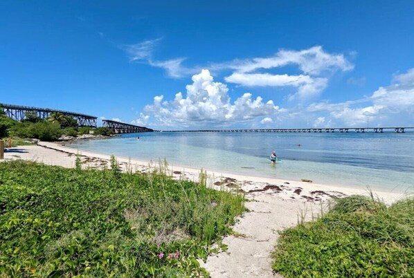 A beach with a clear sky, a person in the water, and a bridge in the background.
