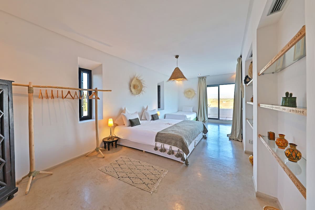 A bedroom with a bed, bedside table, lamp, and a view of the outdoors.