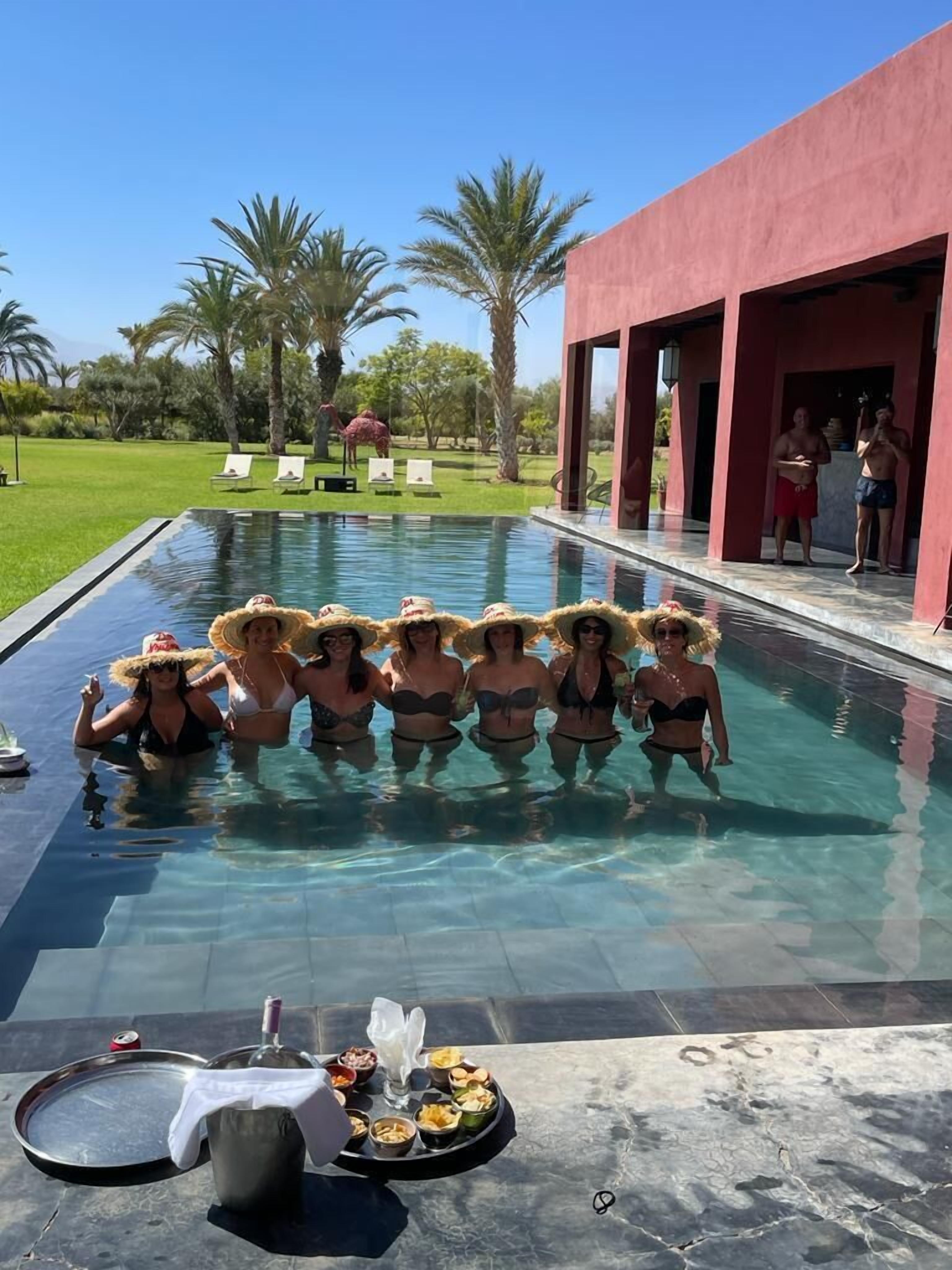 A poolside area with a group of people in swimwear, a dining setup with food and drinks, and a red building in the background.