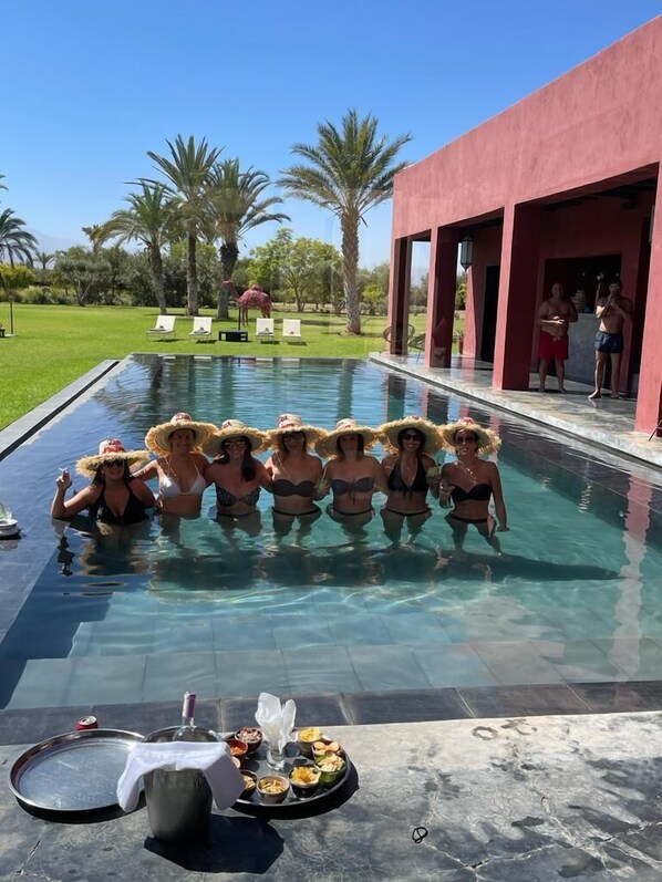 A poolside area with a group of people in swimwear, a dining setup with food and drinks, and a red building in the background. - Dar Noujoum (Ghmate)