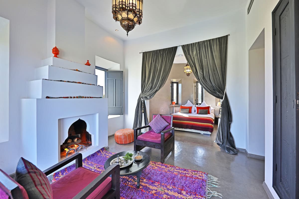 A modern hotel room with a fireplace, a large bed, a seating area with a coffee table, and a view of the city.